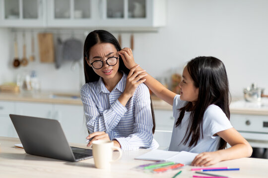 A mother is trying to focus on her work at home on a laptop while her daughter playfully interrupts her. The setting is a cozy kitchen during quarantine, filled with homeschool materials.