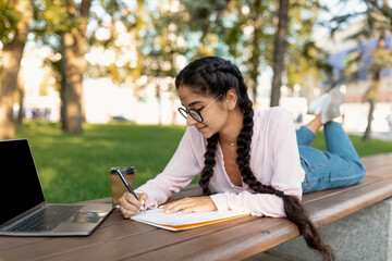 Obraz premium A young woman relaxes on a bench in a park, writing in her notepad while her laptop rests nearby. She enjoys a warm cup of coffee under the shade of trees.