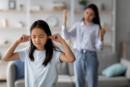 In a home setting, a young Asian girl covers her ears, not wanting to hear her mother’s scolding. The mother expresses frustration, highlighting a family argument.