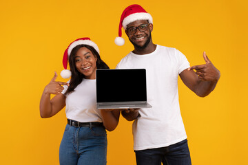 In a lively yellow studio, a cheerful couple points to a blank laptop screen while wearing festive Santa hats. They smile at the camera, promoting a Christmas offer in an engaging way.