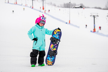 Smiling child in colorful snowboard gear standing on a snowy slope, holding a snowboard . Winter sports .