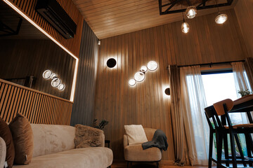 A wood clad living room shows a beige sofa, curved armchair with throws, and cascading circular wall sconces reflected in a backlit mirror, lit by evening pendant bulbs.