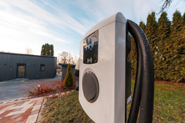 A white wall mounted EV charger with coiled black cable sits by a dark outbuilding, port cover...