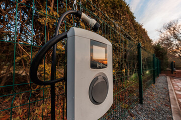 A wall mounted EV charger with coiled cable and Type 2 connector hangs on a green metal fence beside gravel and pavers. Low angle early evening light reflects on the screen. © True Pixel Art