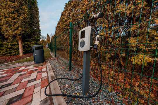 A Level 2 EV charger stands on gravel by a green metal fence, cable holstered, near patterned pavers and a trash bin in a small lot, with warm light and long shadows.