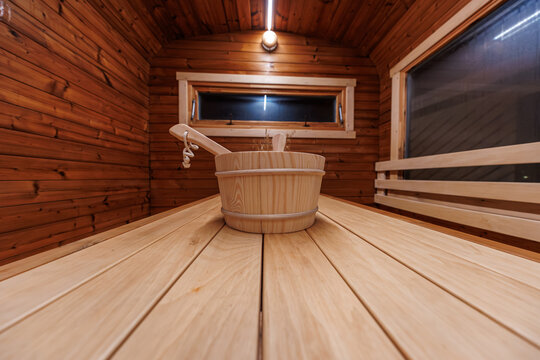 A wooden bucket and long ladle rest on wide bench planks inside a cedar barrel sauna, warm light glows, a narrow window reads dark, low angle shows curved walls.