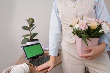 Businesswoman celebrating International Women's Day, holding flowers while standing behind an office desk. Clean desk layout with tablet displaying green screen for content replacement.