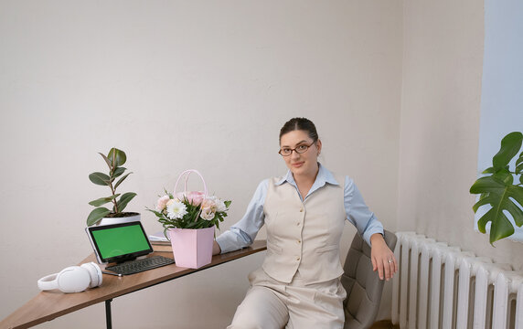 Beige-suited businesswoman holding a floral gift bouquet for March 8 in a modern workspace. Modern workspace with a tablet showing a green screen for design or editing. - Powered by Adobe