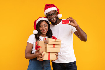 Joyful African American couple stands against a bright yellow background, wearing Santa hats while holding Christmas gift boxes and a credit card. They are smiling and celebrating the holiday spirit.