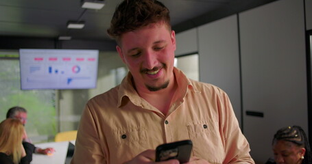 Close-up of young businessman smiling while reading message on smartphone, colleagues in background...