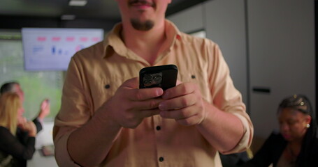 Close-up of young businessman smiling while reading message on smartphone, colleagues in background...