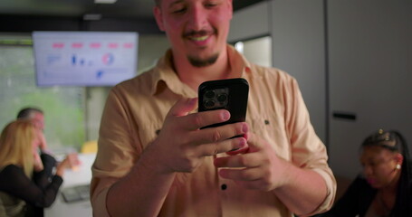 Close-up of young businessman smiling while reading message on smartphone, colleagues in background focused on laptops during meeting in office