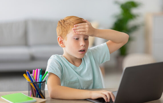A ginger boy is sitting at a table with stationery in front of a laptop. He touches his forehead, looking stressed and unwell as he tries to complete his homework at home.