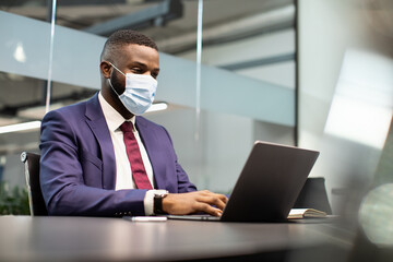 Successful young black businessman in expensive blue suit and face mask working on laptop at office while COVID-19 pandemic, typing on computer keyboard, sending emails, copy space