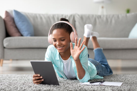 A joyful African American teen girl studies at home, lying on the floor. She uses a tablet for an online call and waves her hand while wearing headphones, enjoying the new normal of remote learning.