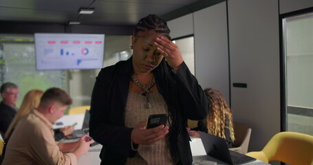 Concerned African American businesswoman looking at smartphone during office meeting, touching forehead while processing information, team in background