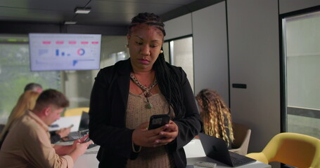 Concerned African American businesswoman looking at smartphone during office meeting, touching forehead while processing information, team in background