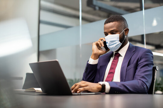 Successful young black businessman in expensive blue suit and face mask working on laptop at office while COVID-19 pandemic, having phone conversation with his personal assistant, copy space