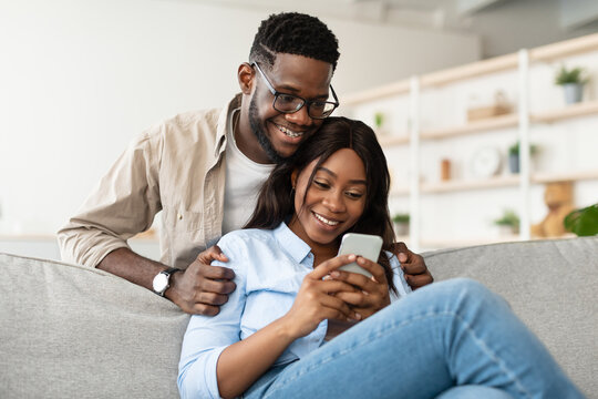 An African American couple enjoys quality time on their couch, smiling and exploring social media together on a smartphone. The man embraces the woman, highlighting their connection. - Powered by Adobe