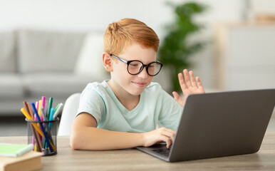 Redhead preteen boy wearing glasses happily engages in a video call with his tutor at home. He sits at a table, smiling and waving while using a laptop. Learning moment captured in a cozy environment.