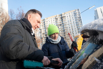 A father and teenage son brush snow off their car. Winter season.