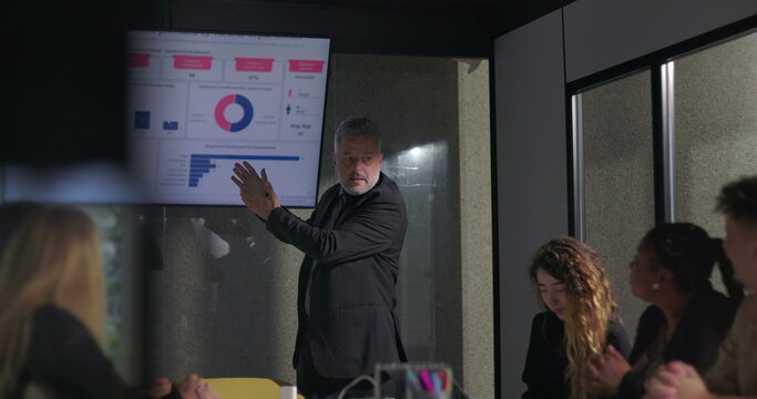 Confident business leader delivering passionate presentation to engaged colleagues during corporate meeting with data charts on screen