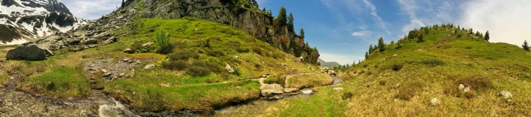 Panoramic alpine landscape with green meadows, rocks, and clear stream under blue sky