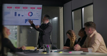 Confident business leader delivering passionate presentation to engaged colleagues during corporate meeting with data charts on screen