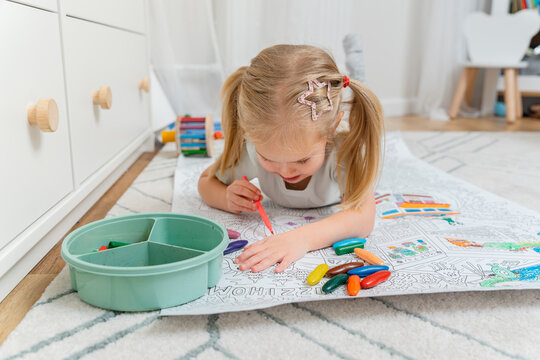 A little girl lies on the floor and paints a coloring book with pencils