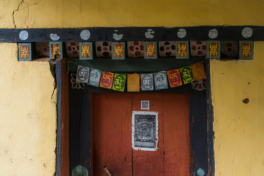 View of a weathered orange door beneath vibrant, colorful Buddhist prayer flags and ornate painted wooden beams on a yellow wall, Paro, Bhutan.