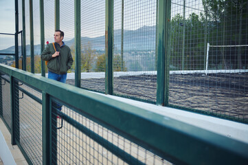 Fototapeta premium Man With Backpack Stands By Fenced Sports Field Gazing Toward Distant Hills