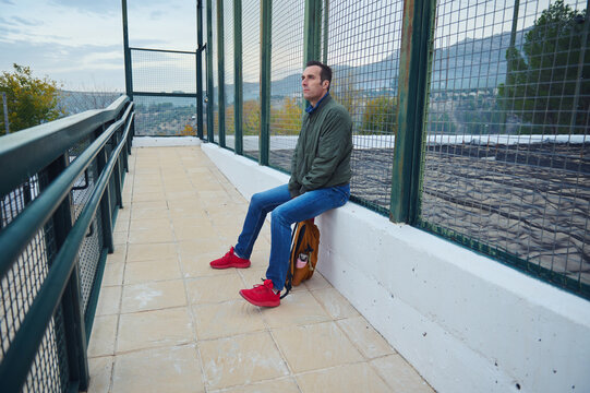 Man Sits On Outdoor Balcony Bench By Fence With Backpack And Red Shoes