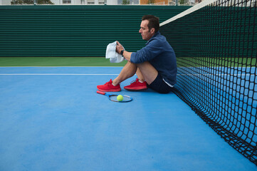 Man Resting on Blue Tennis Court After Match With Ball and Racket