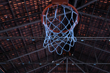 Close-up of a basketball hoop and net in an indoor court. The blurred background shows the roof structure, creating a sports and recreation atmosphere.
