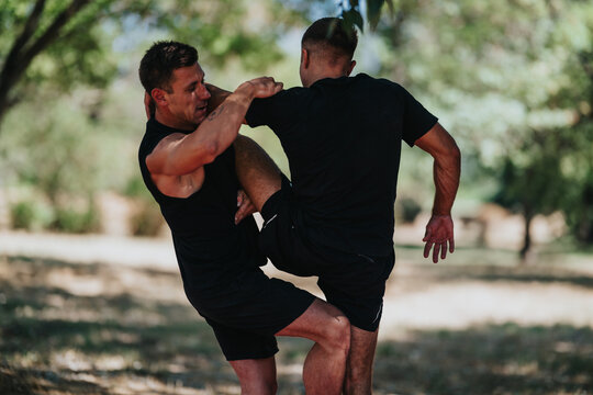 Two athletic men in black shirts practice wrestling and grappling outdoors in a sunny park. A dynamic training scene emphasizing fitness, teamwork, and combat technique.
