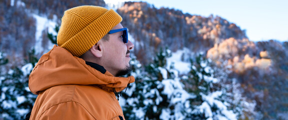Man wearing orange jacket and yellow beanie against snowy mountains and trees , enjoying the tranquility of nature and beauty of outdoors in winter , copy space