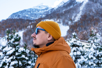 Smiling man traveler wearing sunglasses,orange jacket and yellow beanie against snowy mountains and trees , enjoying the tranquility of nature and beauty of outdoors in winter