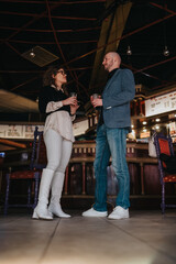 Fototapeta premium Two professionals, a man and a woman, stand facing each other in a dimly lit bar, holding drinks and engaging in a friendly, professional conversation that hints at partnership and networking.