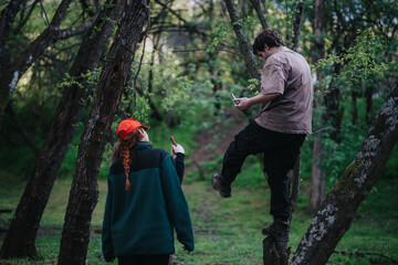 Two people pause on a tree and a path in a green forest, looking at their phones. A candid outdoor scene that conveys friendship, curiosity, and modern connectivity.