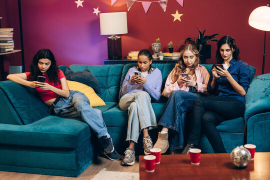 Young women using smartphones on sofa at party, showing digital addiction and social disengagement
