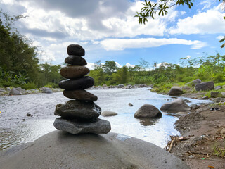 	A perfectly balanced stack of smooth river stones, or cairn, stands tall on a larger rock by a flowing river under a bright, cloudy sky, surrounded by lush green foliage and trees, symbolizing peace 