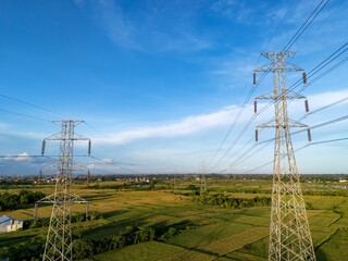 High voltage electrical transmission towers (pylons) carrying power lines over green rice fields and residential areas under a blue sky. Energy and nature contrast.