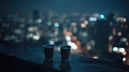 Two disposable coffee cups sit on a rooftop edge as city lights blur into bokeh in the night. Concept Rooftop night photography, City lights bokeh, Coffee cups on rooftop, Urban nightscape