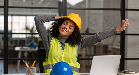 Happy female construction engineer stretching arms at desk in modern office. Relaxed woman worker with yellow hard hat and safety vest taking a break. Work satisfaction concept