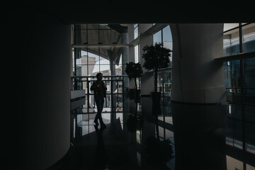 A lone man strides through a dim, contemporary office lobby featuring floor-to-ceiling glass, polished surfaces, and potted trees, creating dramatic reflections and a quiet, introspective atmosphere.
