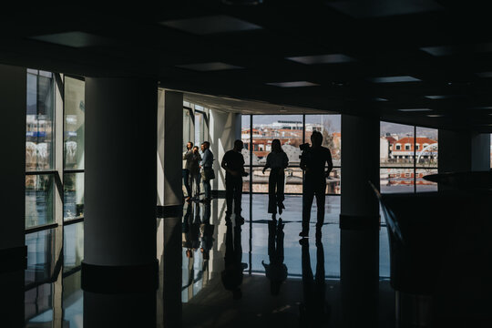 A dark, reflective lobby scene shows several silhouettes gathered near floor-to-ceiling windows, creating dramatic shadows as city houses glow outside.