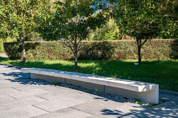 Concrete bench under trees in a park on a sunny day with green grass and hedges in the background