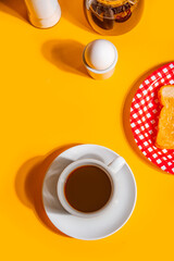 A cozy retro scene featuring a white coffee cup filled with black coffee on a yellow background, accompanied by a glass coffee pot, toasts, egg and a cream pitcher.