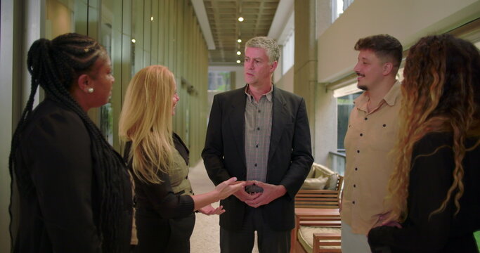 Corporate manager speaking to diverse team in modern office hallway, group of professionals engaged in workplace conversation during informal meeting