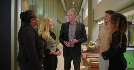 Corporate manager speaking to diverse team in modern office hallway, group of professionals engaged in workplace conversation during informal meeting
